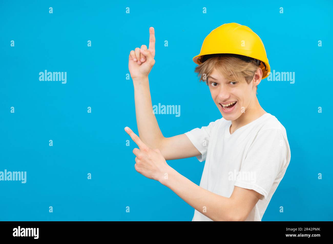 A teenage boy in a yellow helmet is looking at the camera and points fingers up, standing in a studio on a blue background. The concept of constructio Stock Photo