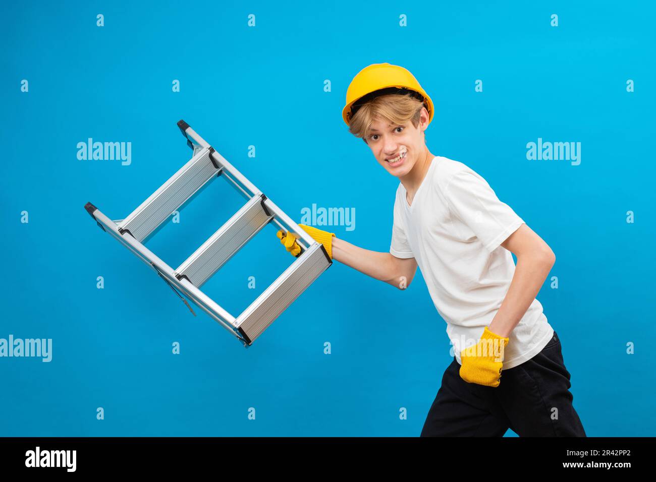 Happy repairman teen in white t-shirt and gloves with yellow helmet in head, holding ladder in hands and running in studio on blue background Stock Photo