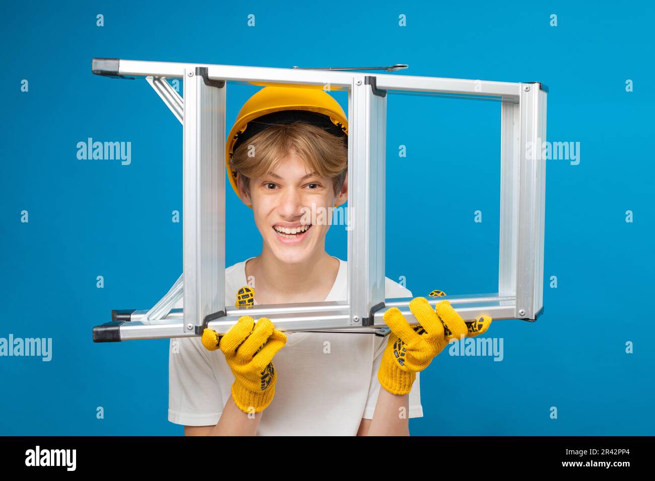 Happy repairman teen in white t-shirt and gloves with yellow helmet in head, holding ladder in hands standing in studio on blue background. Stock Photo