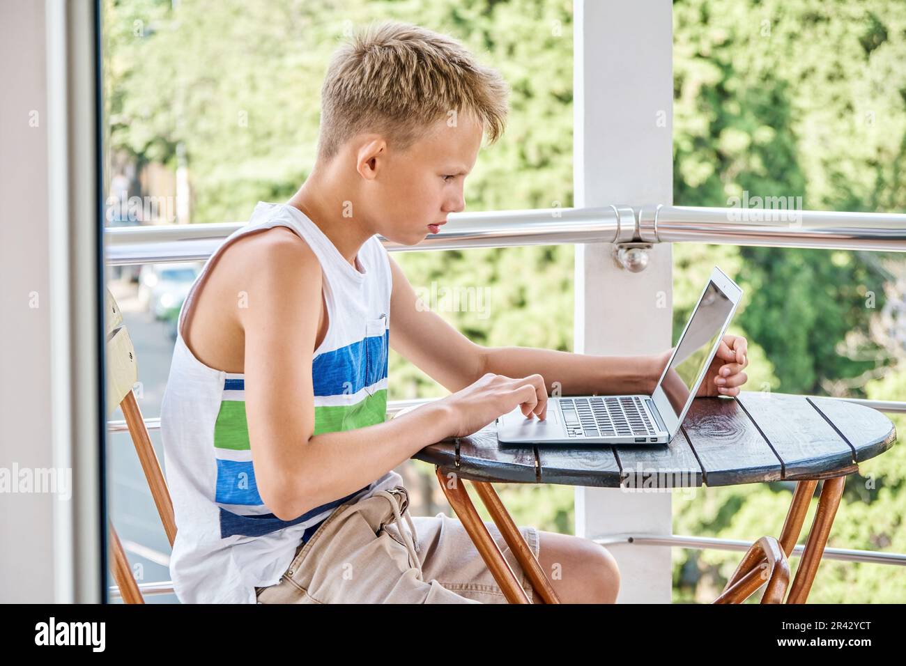 Preteen boy sitting on hotel balcony and surfing the internet on grey laptop. Schoolboy enjoys spending summer holidays and playing games Stock Photo