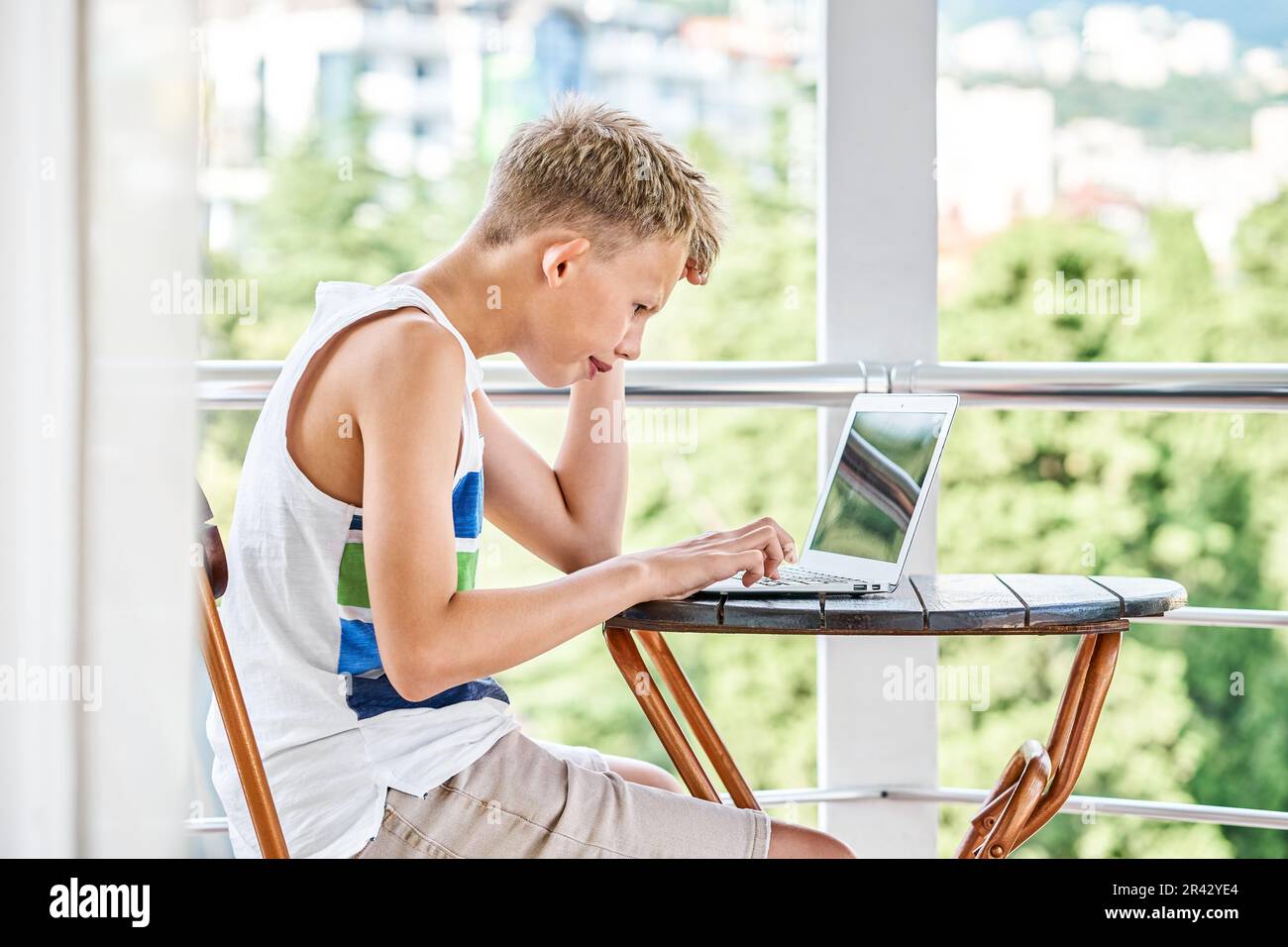 Preteen boy sitting on hotel balcony and surfing the internet on grey laptop. Schoolboy enjoys spending summer holidays and playing games Stock Photo