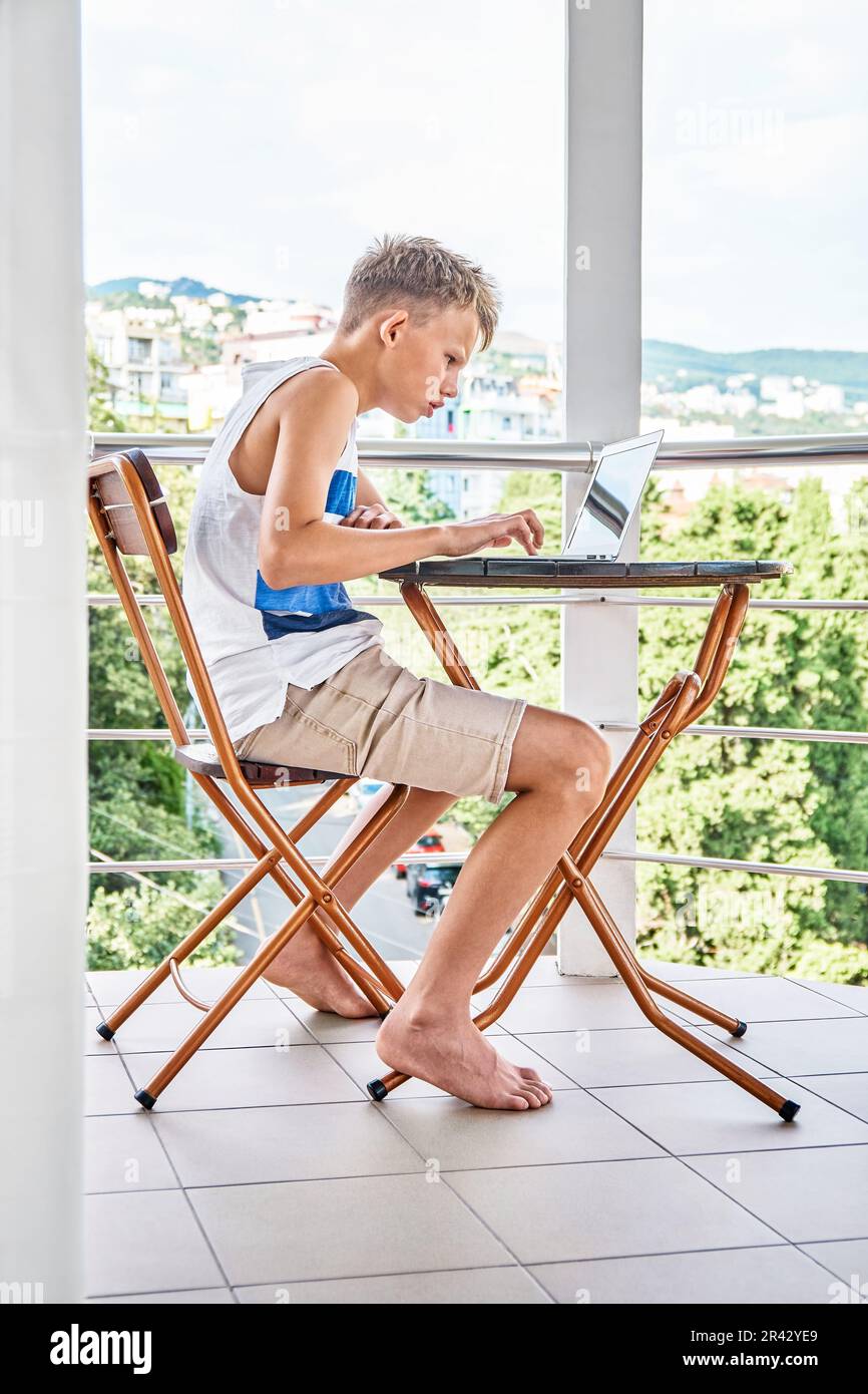 Preteen boy sitting on hotel balcony and surfing the internet on grey laptop. Schoolboy enjoys spending summer holidays and playing games Stock Photo