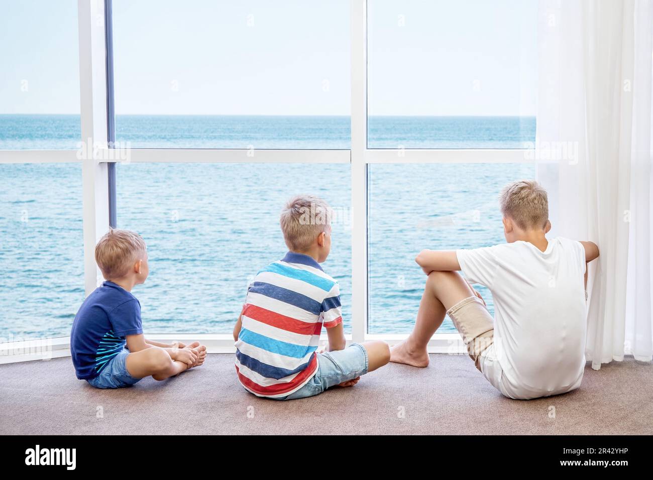 Boys siblings enjoy watching azure sea from panoramic window in hotel room. Blond brothers spend summer holidays at seaside resort together Stock Photo