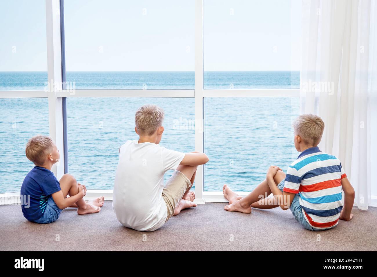 Boys siblings enjoy watching azure sea from panoramic window in hotel room. Blond brothers spend summer holidays at seaside resort together Stock Photo