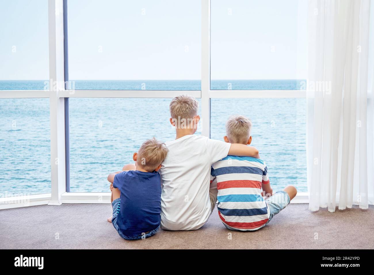 Eldest boy hugs younger brothers sitting on floor and looking at azure sea from panoramic window. Siblings enjoy spending summer holidays together Stock Photo