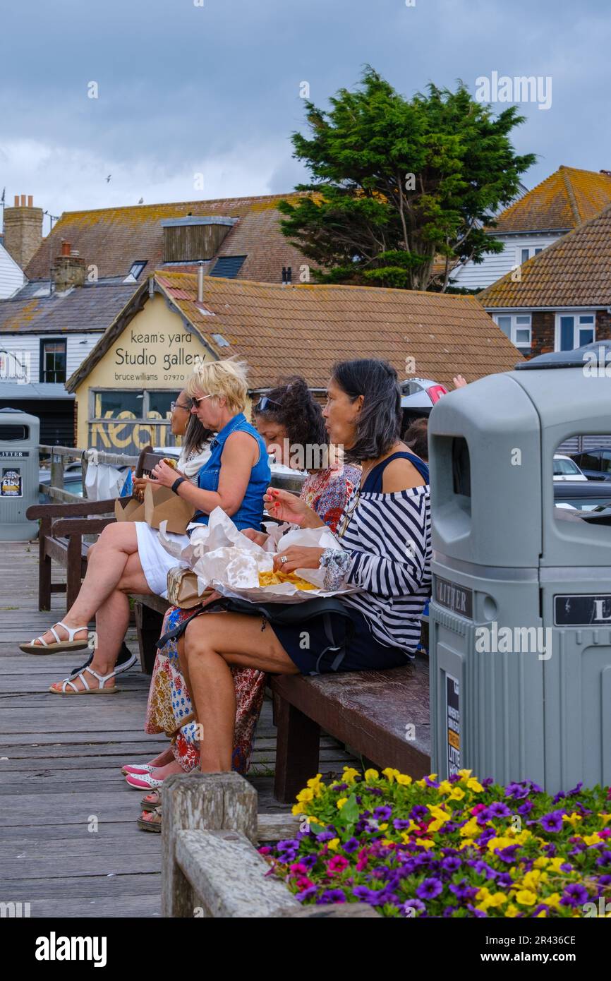 Ladies sitting on a bench at Whitstable beachfront, eating traditional fish and chips. North-east Kent coast, England, UK. Stock Photo