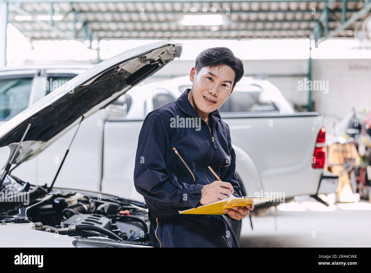 Happy Asian Mechanic young male staff worker working with car engine front hood auto service check tuning engine in garage Stock Photo