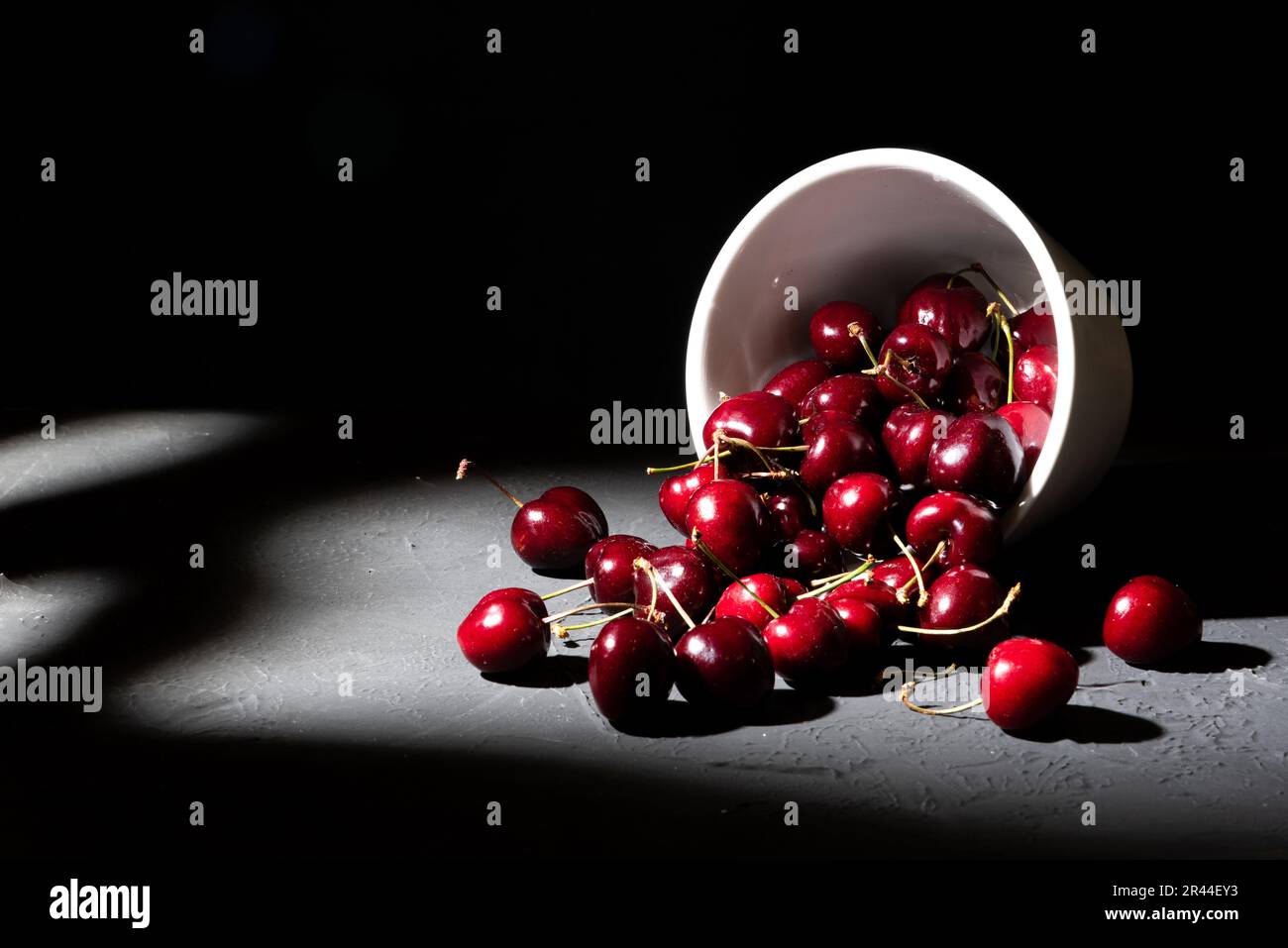 Bowl loaded with red cherries on dark background. Overturned cup spilling rich and healthy cherries Stock Photo