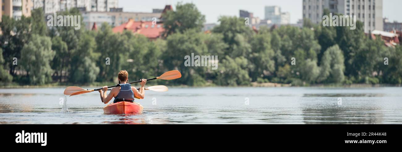 back view of active multiethnic friends in life vests sailing in sportive kayak with paddles on city river with green bank during recreation weekend i Stock Photo