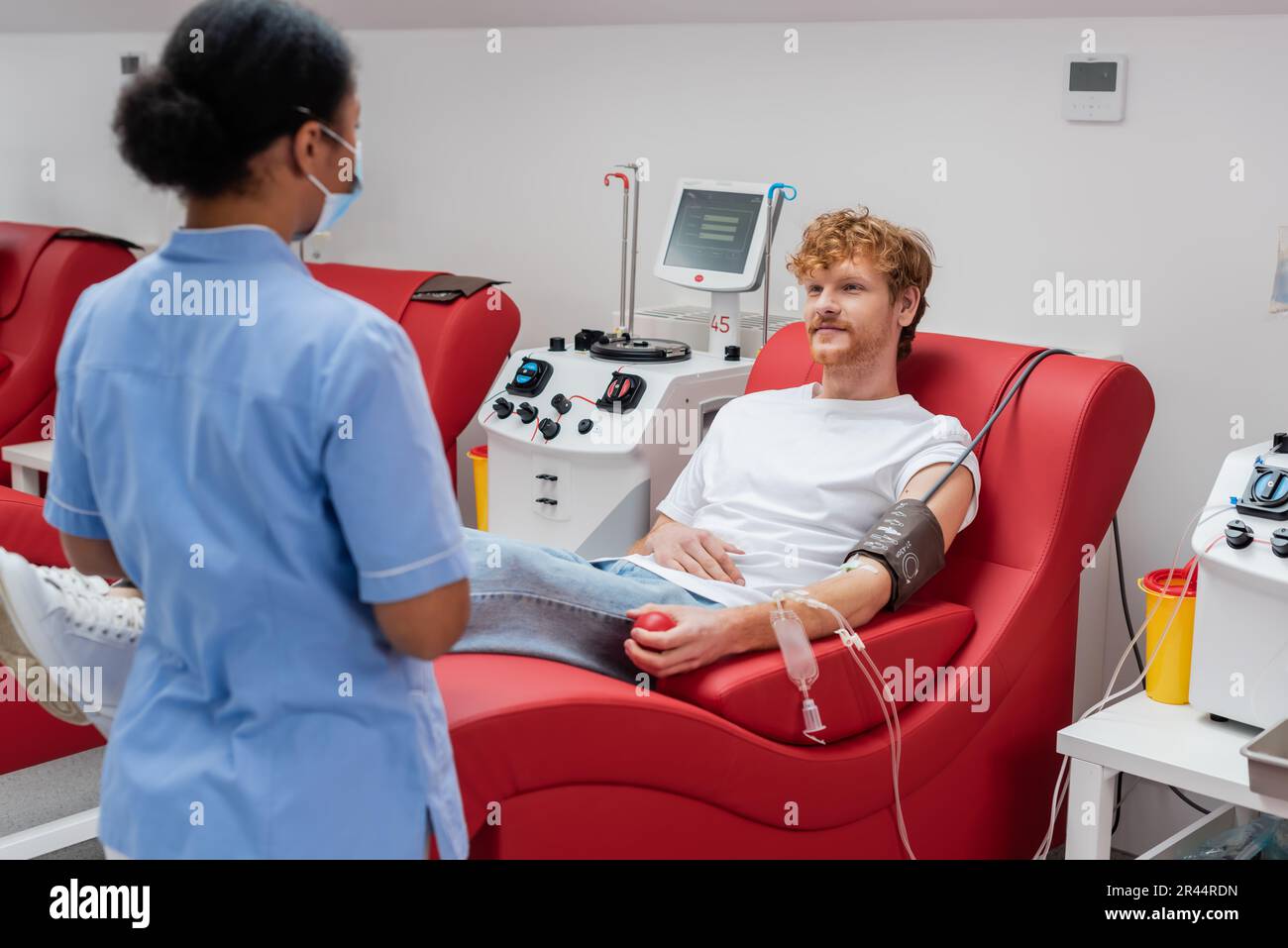 redhead man in blood transfusion set sitting on comfortable chair near automated equipment and multiracial nurse in blue uniform and medical mask in h Stock Photo