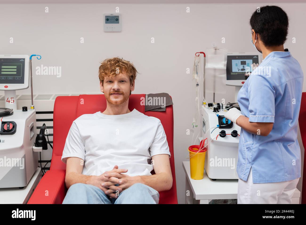 multiracial nurse in blue uniform operating transfusion machine near plastic cup and redhead volunteer sitting on medical chair and looking at camera Stock Photo