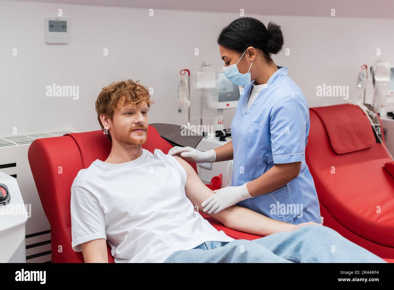 multiracial nurse in medical mask and latex gloves looking at arm of redhead blood donor sitting on comfortable medical chair near transfusion machine Stock Photo