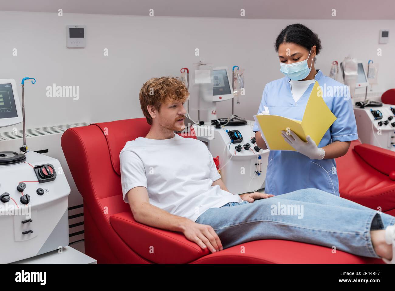 multiracial nurse in medical mask and blue uniform writing in paper folder near transfusion machine and redhead volunteer on medical chair in blood do Stock Photo