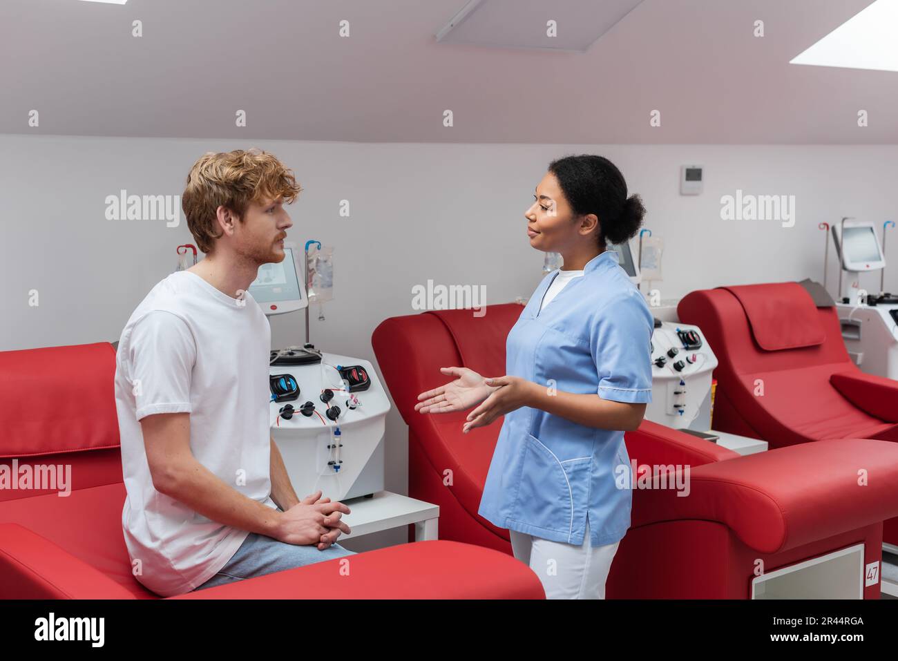 multiracial nurse in blue uniform gesturing and talking to young redhead blood donor sitting on medical chair near transfusion machines in hospital Stock Photo