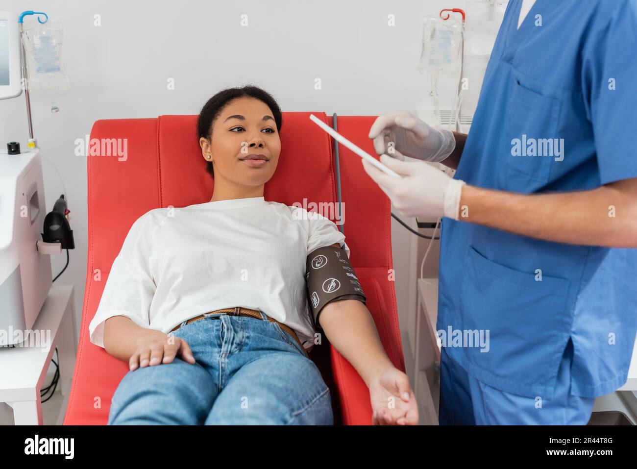 multiracial woman in blood pressure cuff sitting on medical chair near automated transfusion machines and looking at doctor in blue uniform and latex Stock Photo