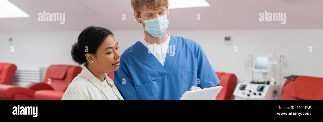 multiracial woman and redhead practitioner in blue uniform and medical mask looking at digital tablet near medical chairs and blood transfusion machin Stock Photo