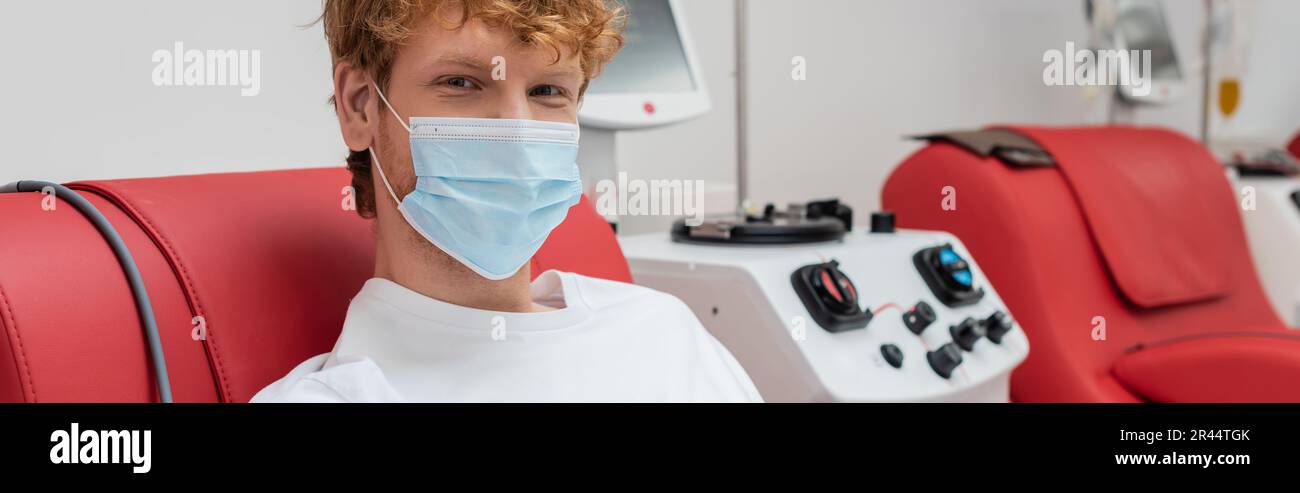 young and redhead volunteer in medical mask, with positive eyes expression, donating blood near contemporary transfusion machine and looking at camera Stock Photo