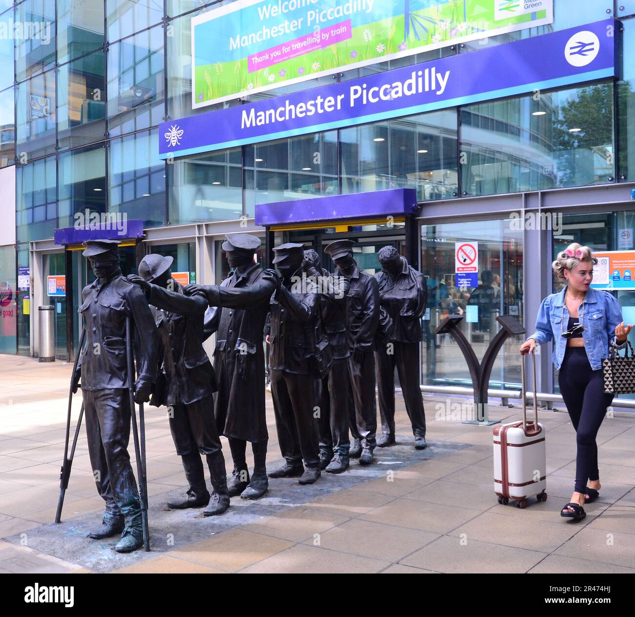 A woman walks past a statue of seven blinded soldiers outside the main entrance of Manchester Piccadilly Railway Station, central Manchester, UK.  Marking the centenary of the end of the First World War, the statue is entitled, 'Victory Over Blindness'. It was commissioned by the military charity Blind Veterans UK. The original sculpture was conceived and designed by artist and sculptor Johanna Domke-Guyot. Stock Photo