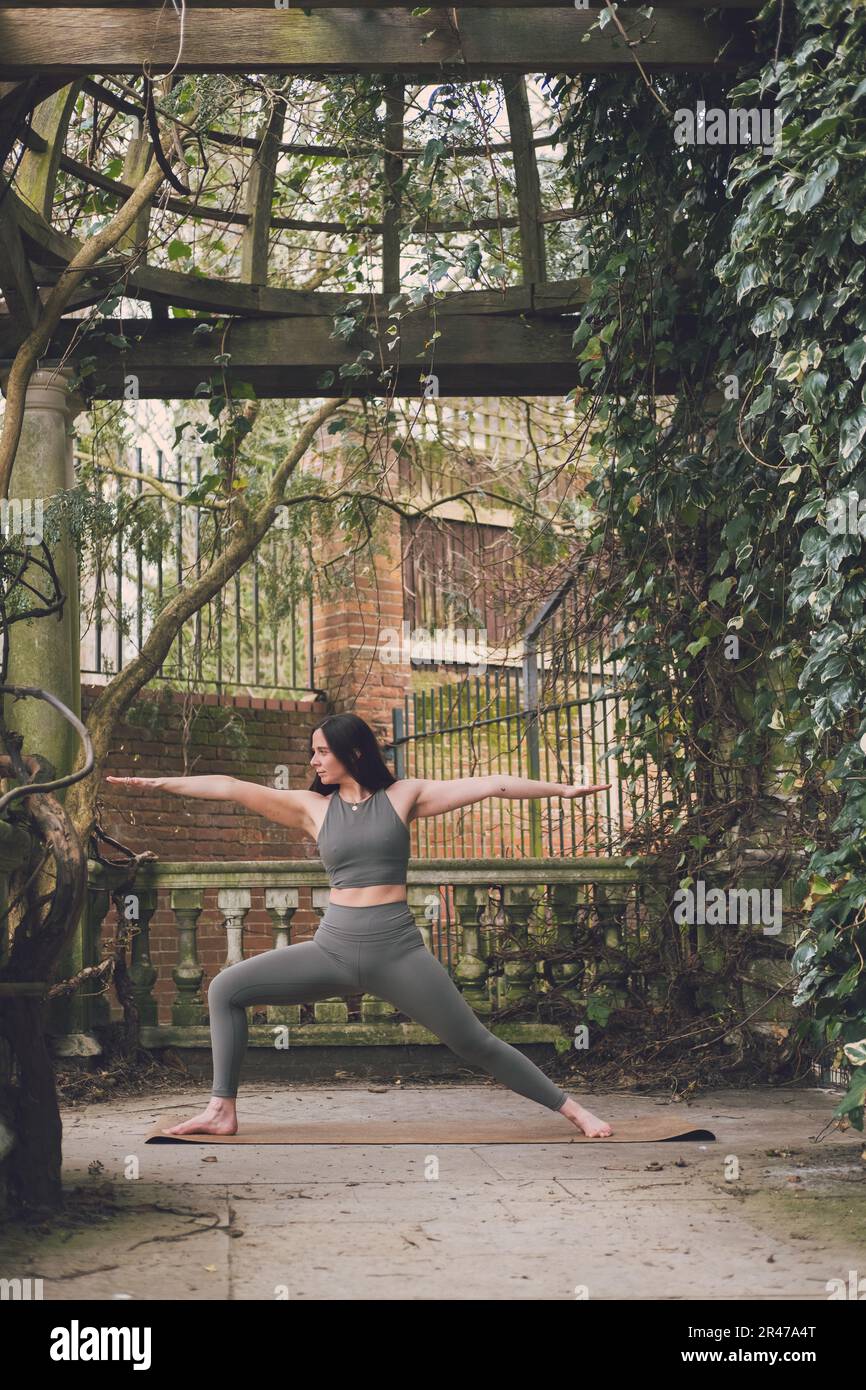 A young adult woman strong new yoga teacher in warrior two pose practicing  in an outside location with columns, wooden beams, plants and leaves. Weari  Stock Photo - Alamy