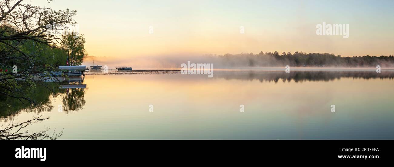Panorama of a calm northern Minnesota lake and fog at dawn during spring with docks and boats along the shoreline Stock Photo
