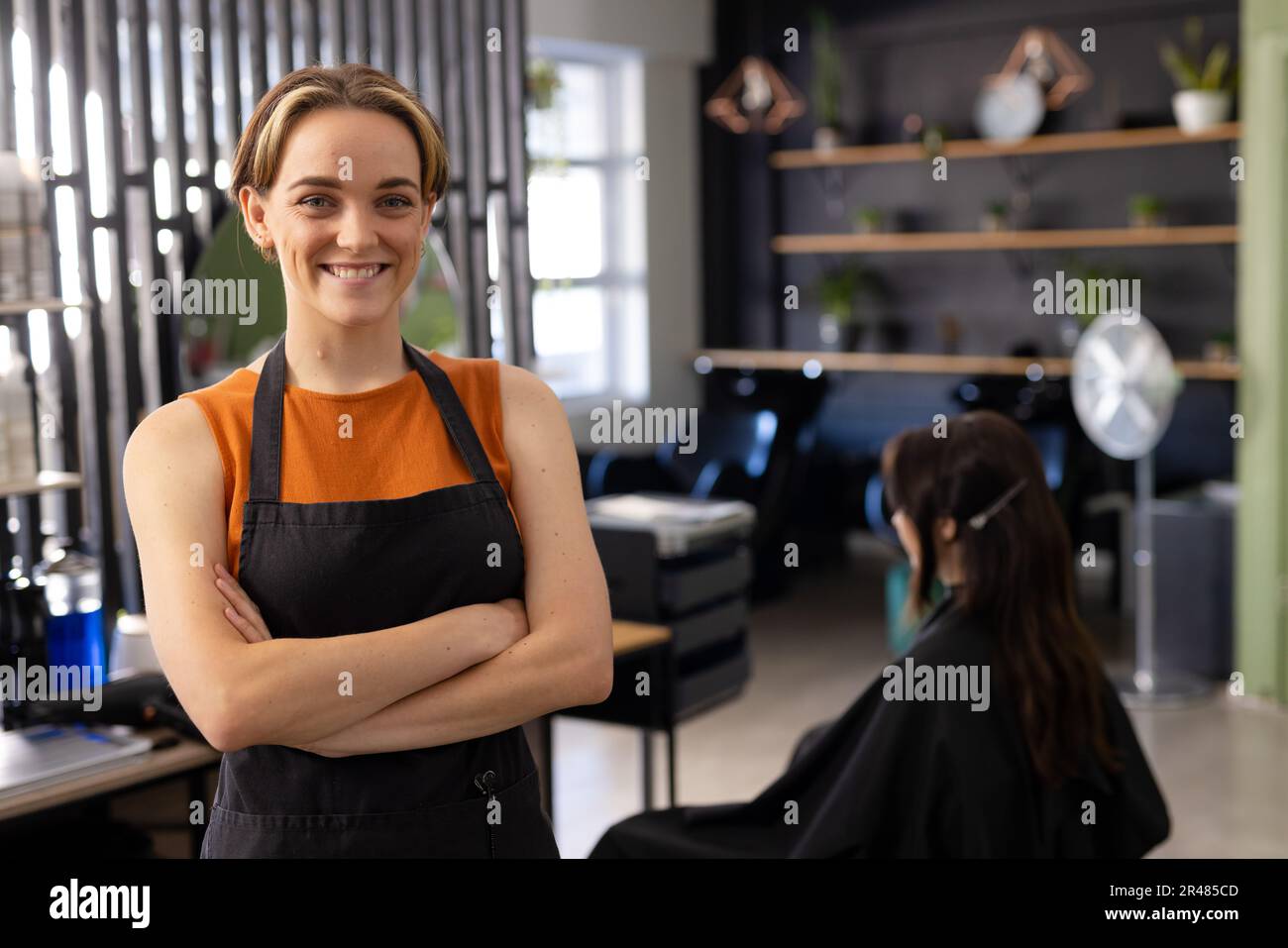 Portrait of smiling caucasian female hairdresser in black apron at hair salon, copy space Stock Photo