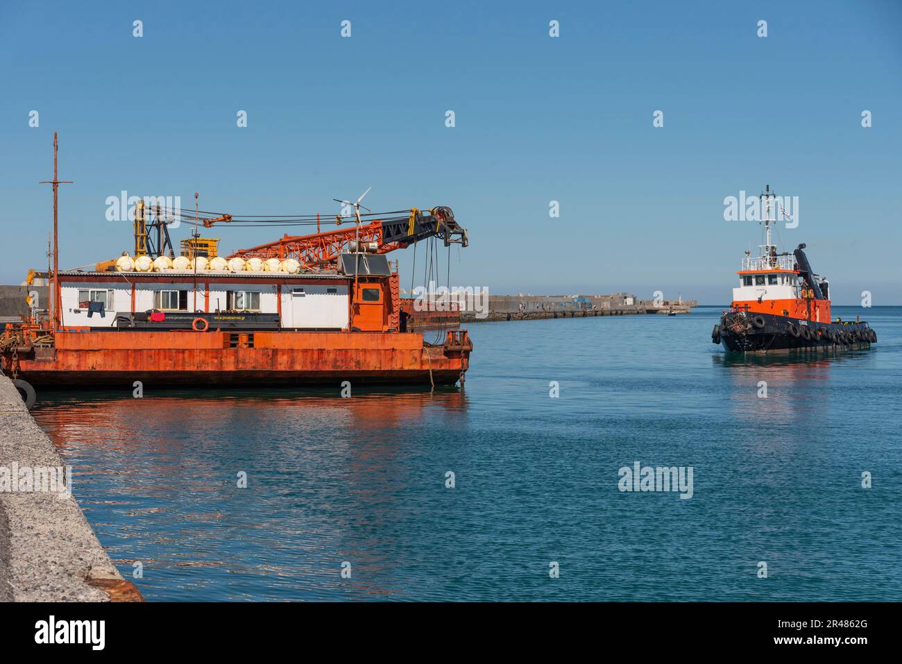 Heraklion, Crete, Greece. 2023. Tug aapproaching a large sea going crane  barge with a crane mounted on deck alongside the harbour. Stock Photo