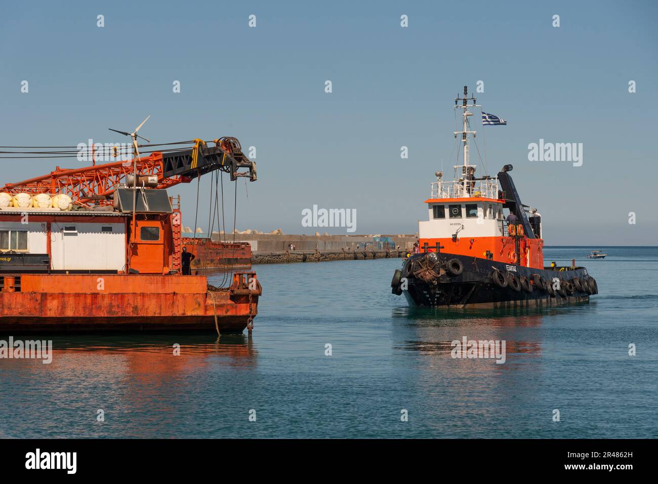Heraklion, Crete, Greece. 2023. Tug approaching a large sea going crane  barge with a crane mounted on deck alongside the harbour. Stock Photo