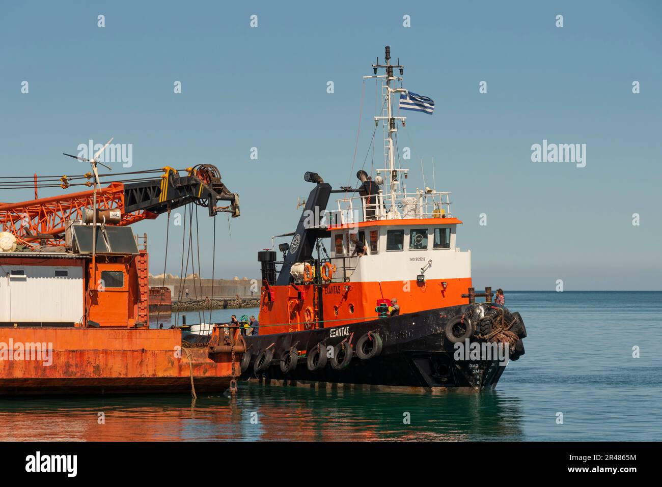 Heraklion, Crete, Greece. 2023. Tug  coming alongside a large sea going crane  barge with a crane mounted on deck alongside the harbour. Stock Photo