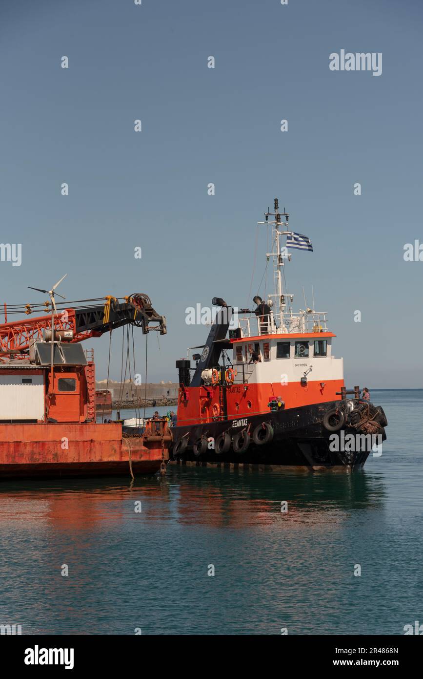 Heraklion, Crete, Greece. 2023. Tug  coming alongside a large sea going crane  barge with a crane mounted on deck alongside the harbour. Stock Photo