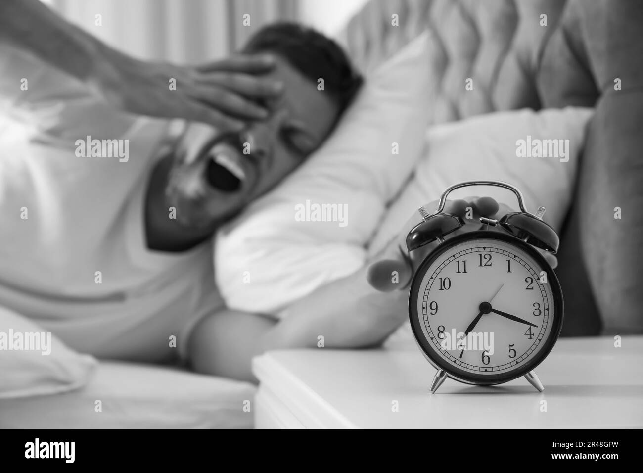 Sleepy man turning off alarm clock at home in morning, selective focus. Black and white photography Stock Photo