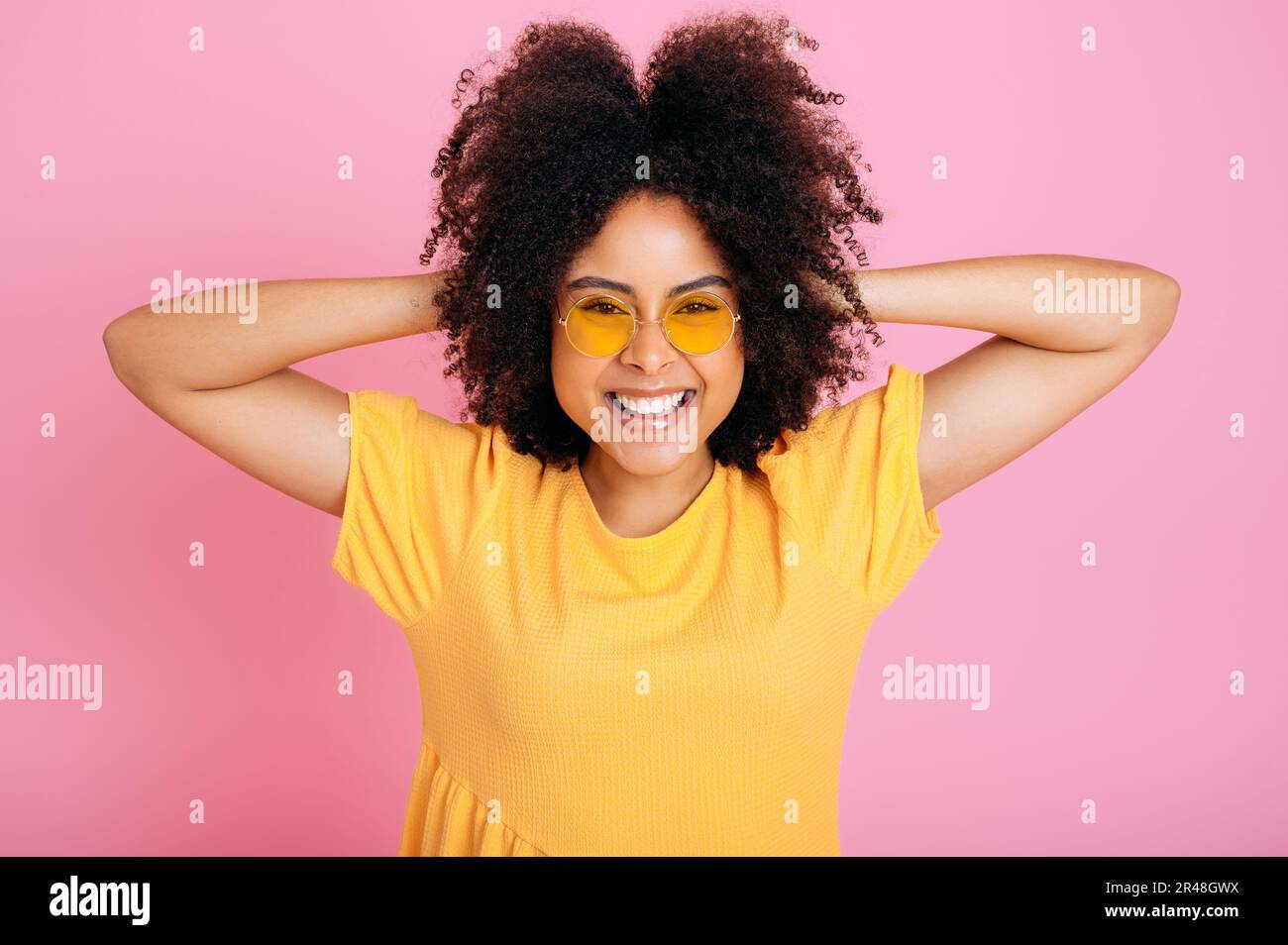 Joyful happy excited brazilian or hispanic curly young woman in trendy orange glasses and sundress, having fun, posing, holding hands on hair, looking at camera, smiling, isolated pink background Stock Photo