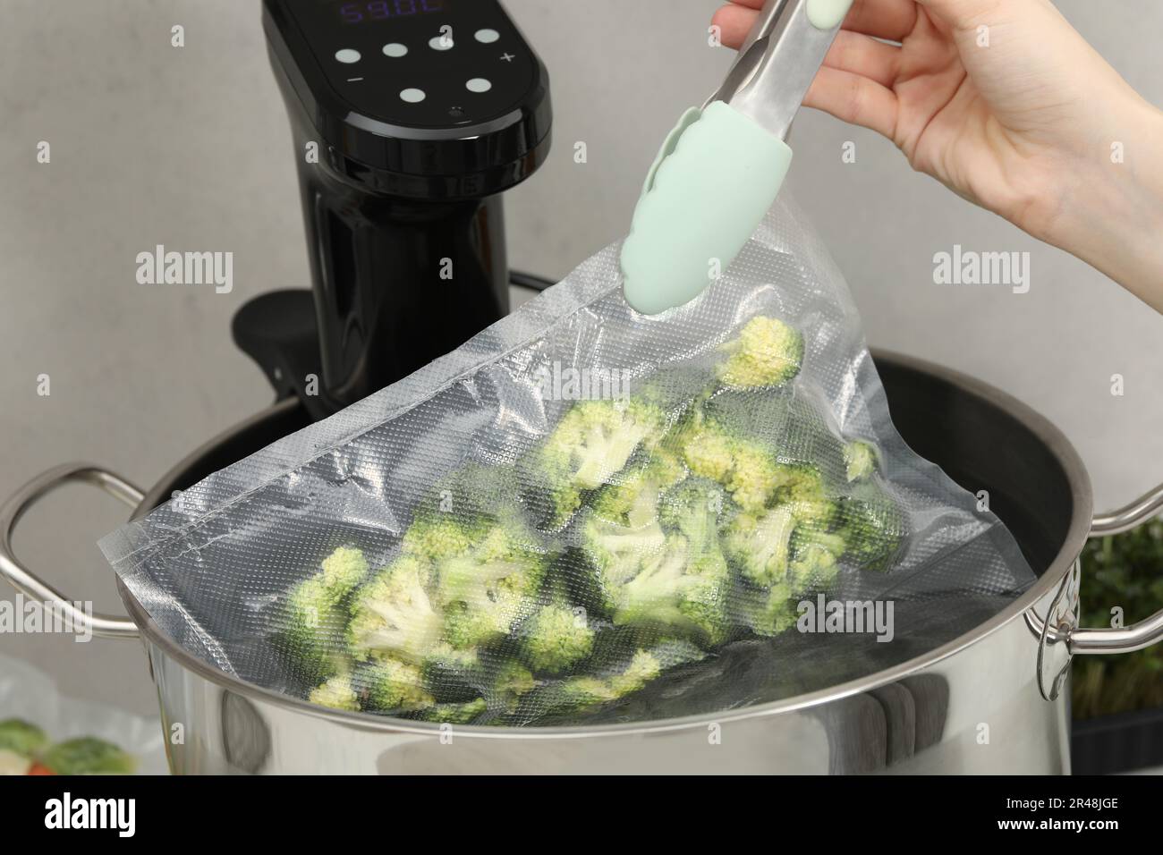 Woman putting vacuum packed broccoli into pot with sous vide cooker, closeup. Thermal immersion