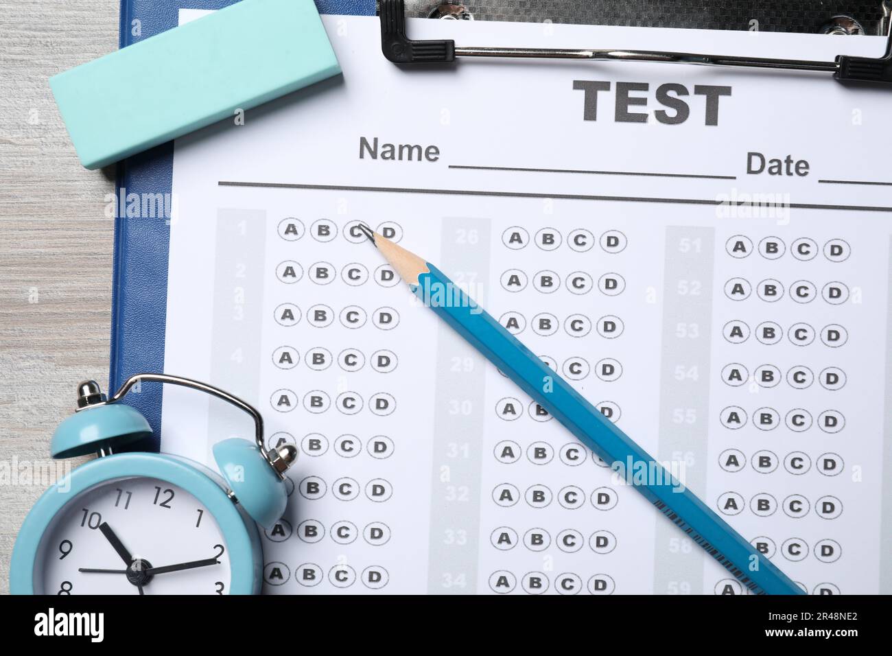 Clipboard with answer sheet, pencil and alarm clock on light wooden