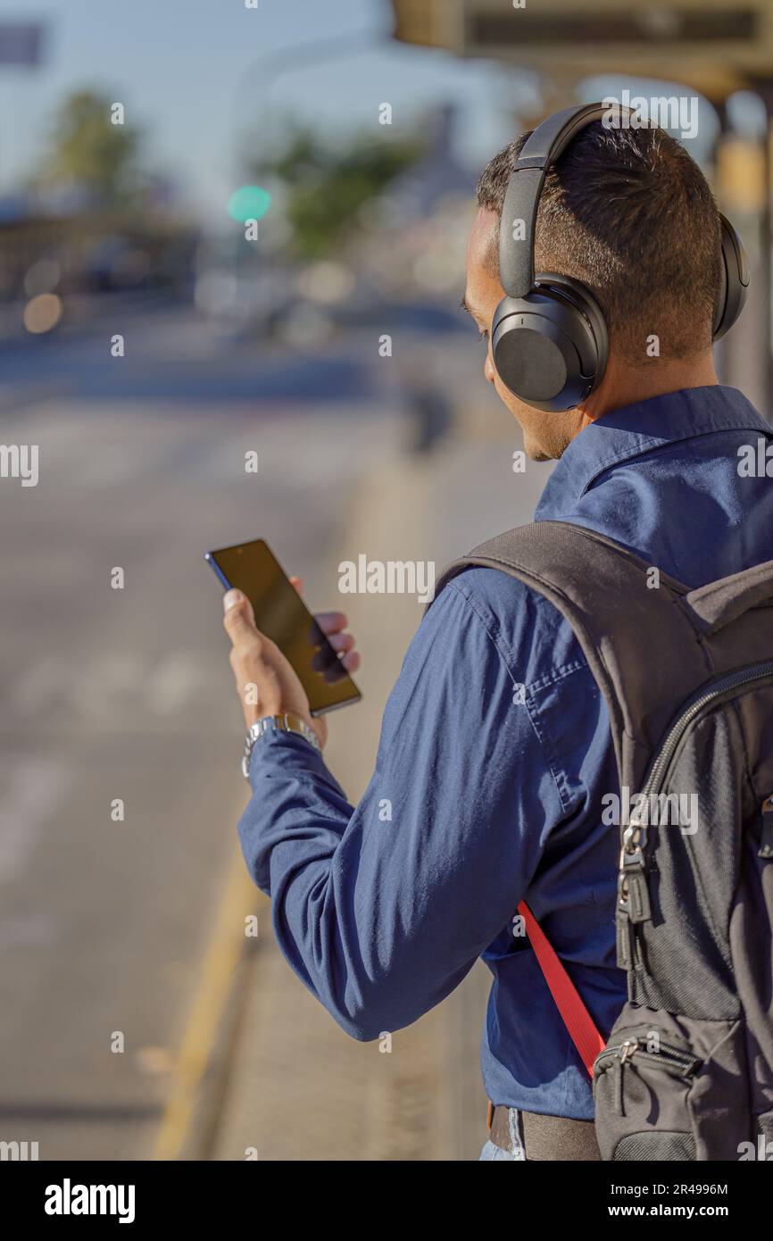 Young latin man with headphones looking at his mobile phone at the bus stop seen from behind. Stock Photo