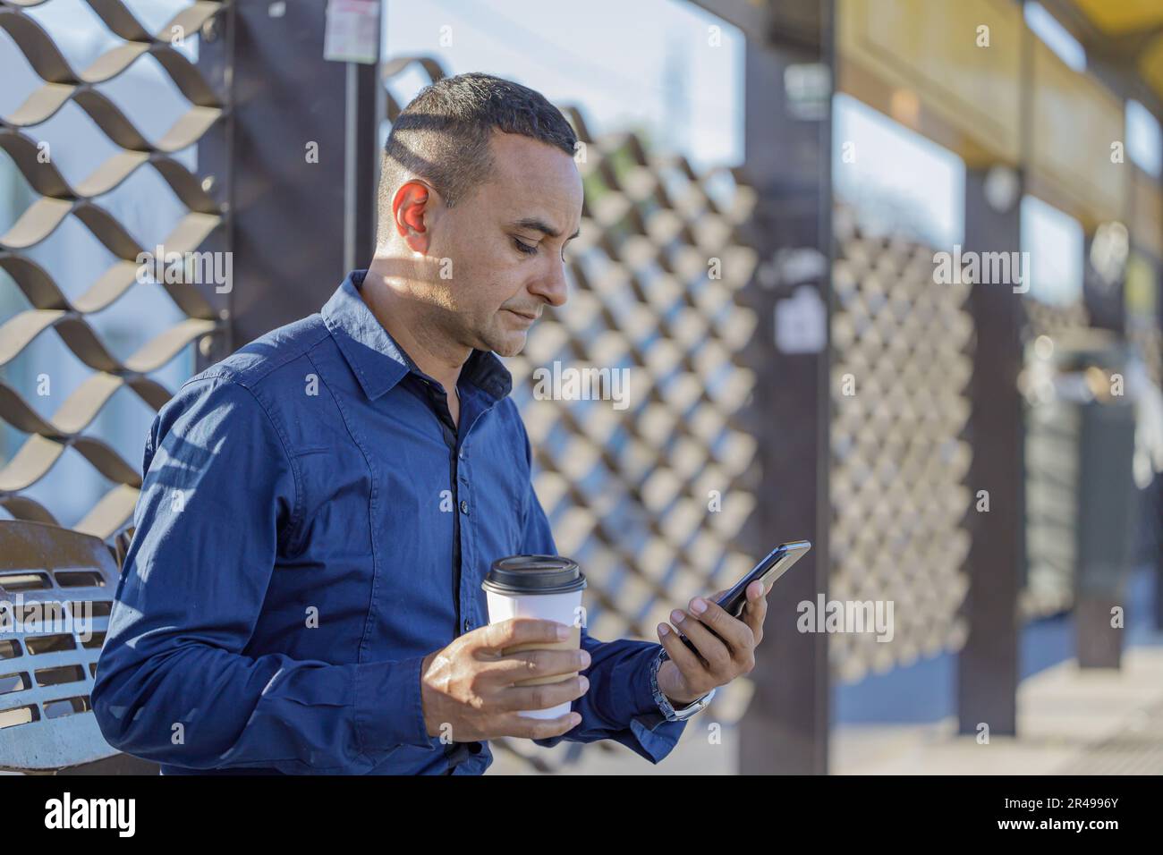 Medium shot of young latin man sitting waiting for the bus looking his mobile phone. Stock Photo