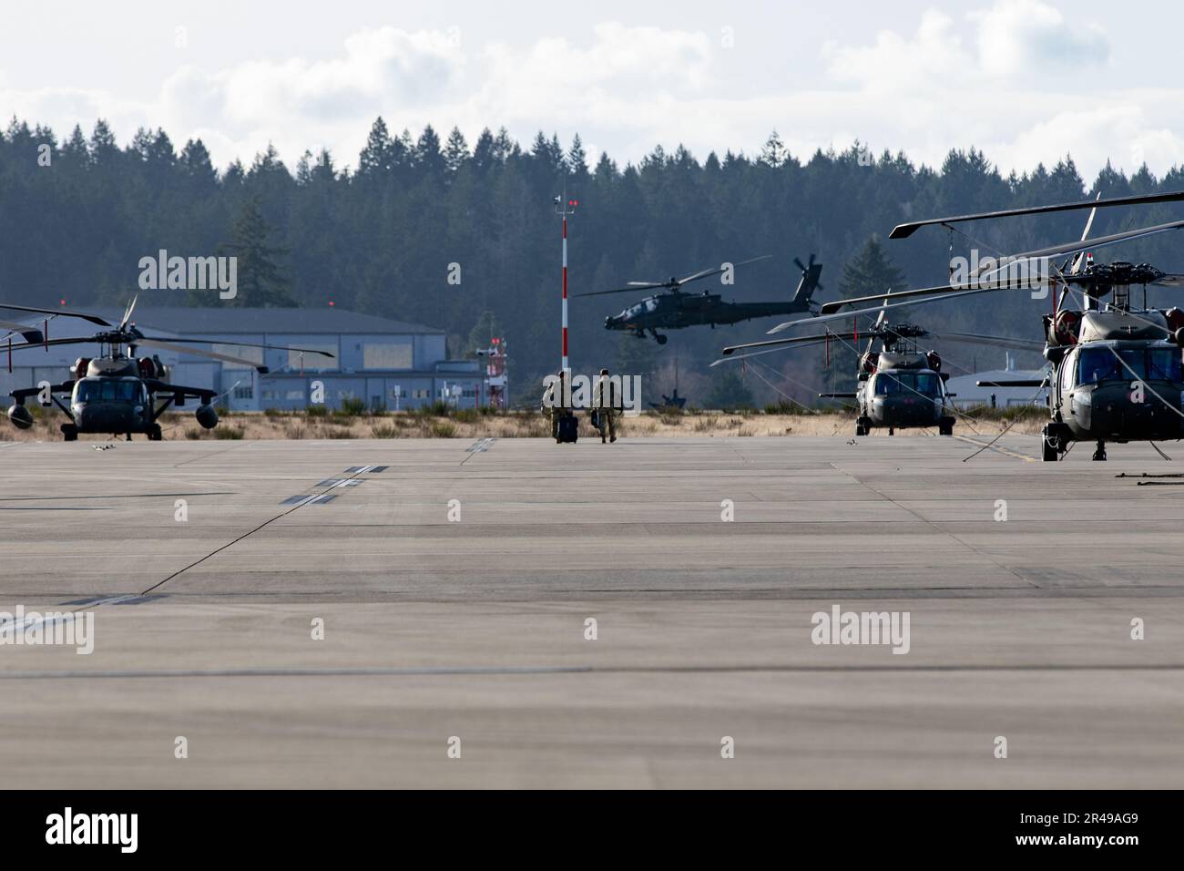 An aircrew assigned to 2-158 Assault Helicopter Battalion, 16th Combat Aviation Brigade walks between a row of parked UH-60M Black Hawk Helicopters at Joint Base Lewis-McChord, Wash. on Feb. 8, 2023. An AH-64E Apache helicopter is flying in the background. Stock Photo