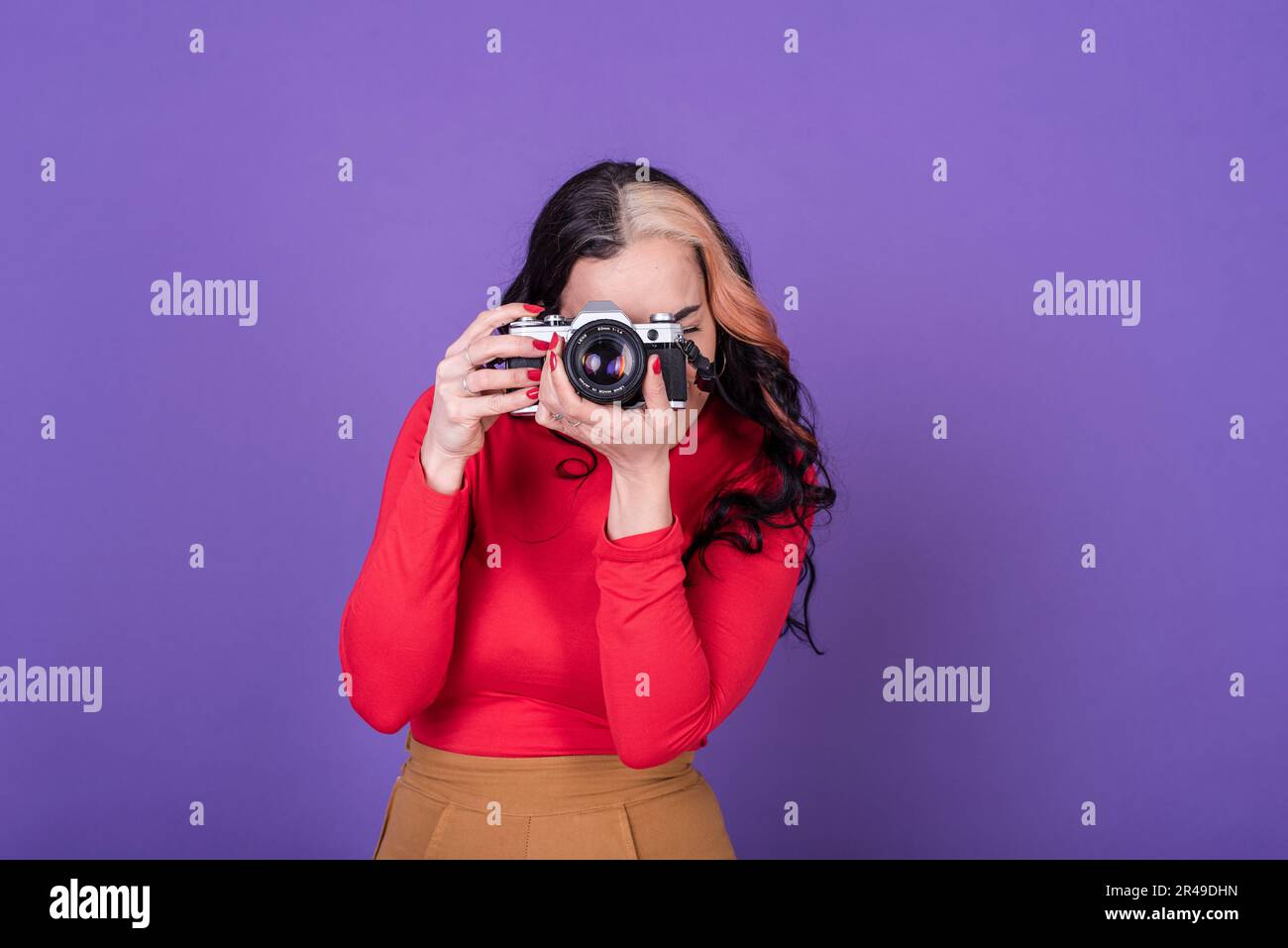 Attractive young lady taking a photo with her film camera over a violet background.  Studio shot. Stock Photo
