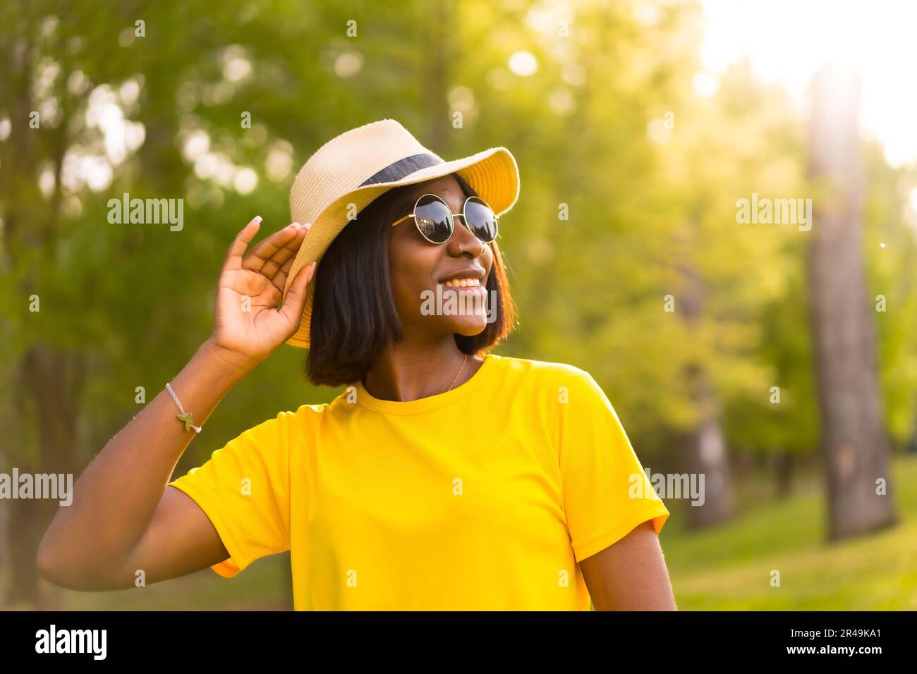 A cheerful black female wearing sunglasses and a hat outdoors in the park on a sunny day Stock Photo