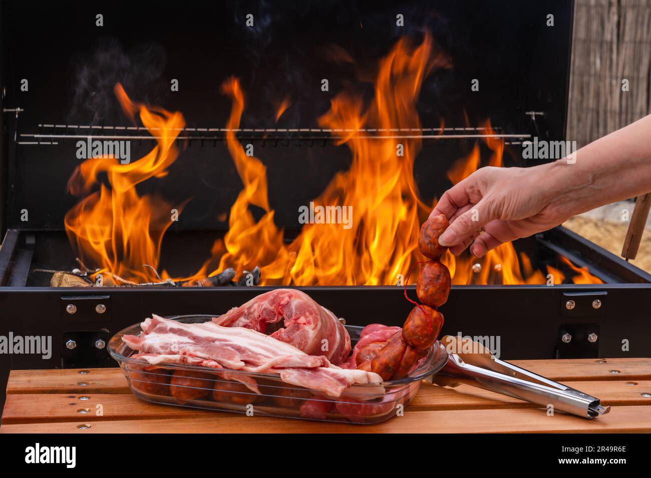 A hand near an array of raw meats and sausage near a burning grill with flames Stock Photo