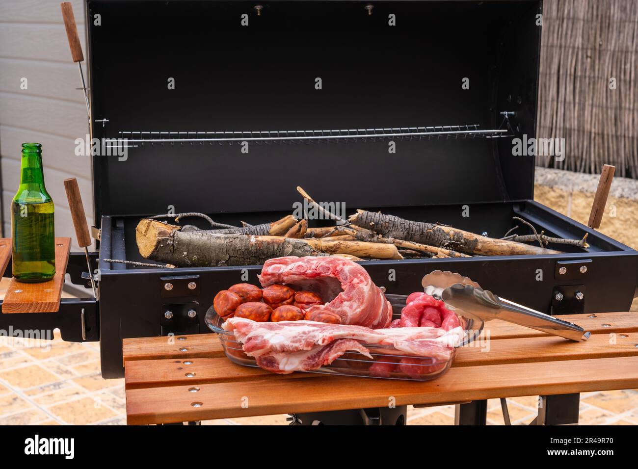 A closeup shot of an array of raw meats near an outdoor grill with wood Stock Photo
