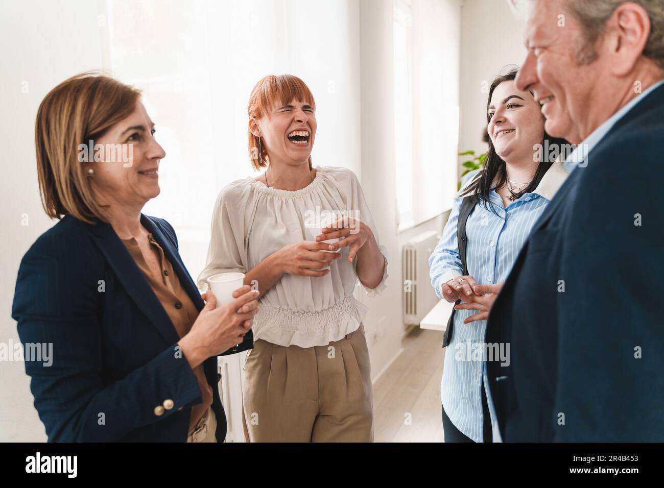 Colleagues of mixed ages and genders taking a break in office, standing near a window, laughing and chatting. Focus is on young redhead woman laughing Stock Photo