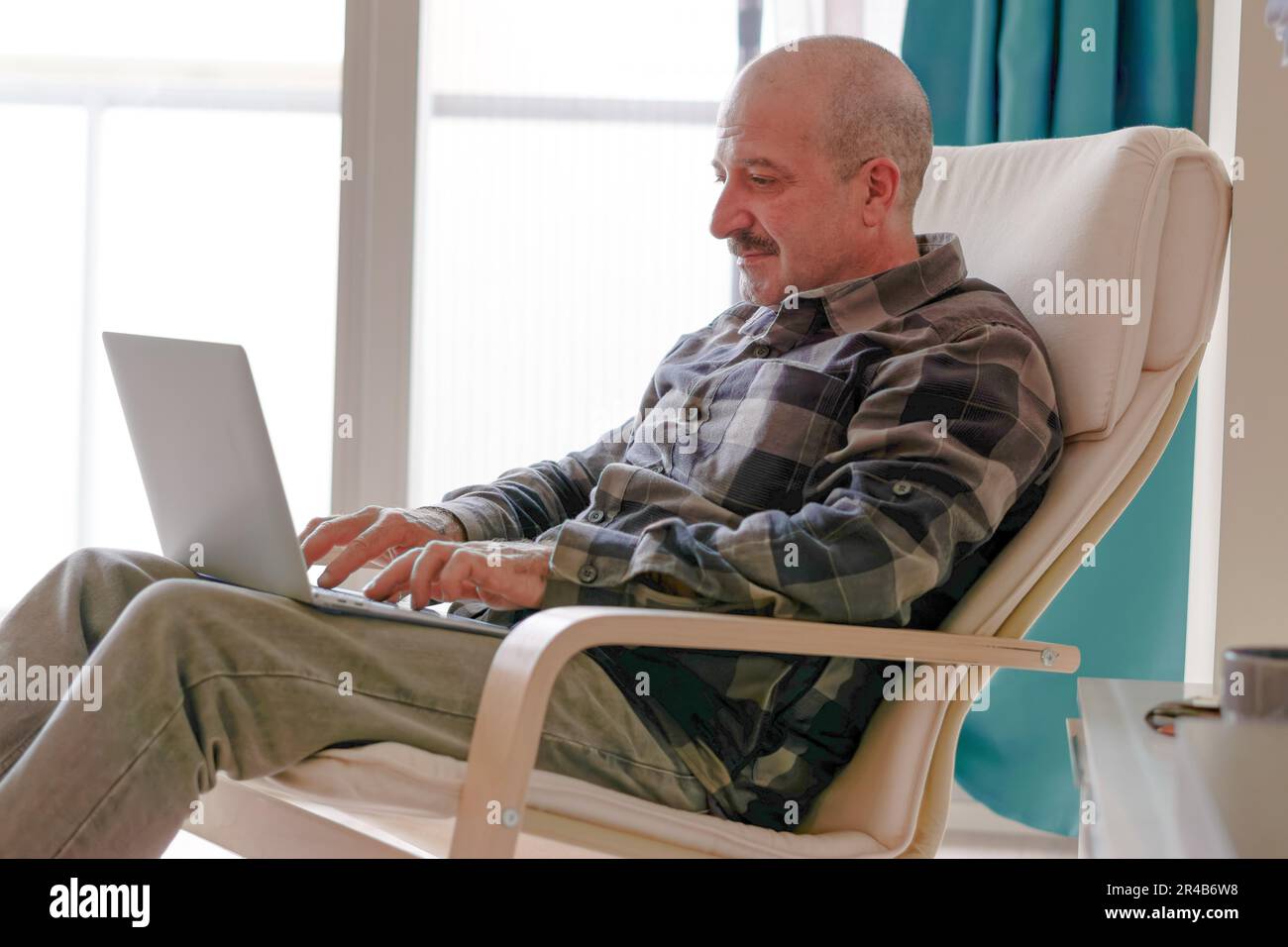 Man with moustache, plaid shirt and jeans sitting in an armchair in his living room using his laptop computer Stock Photo