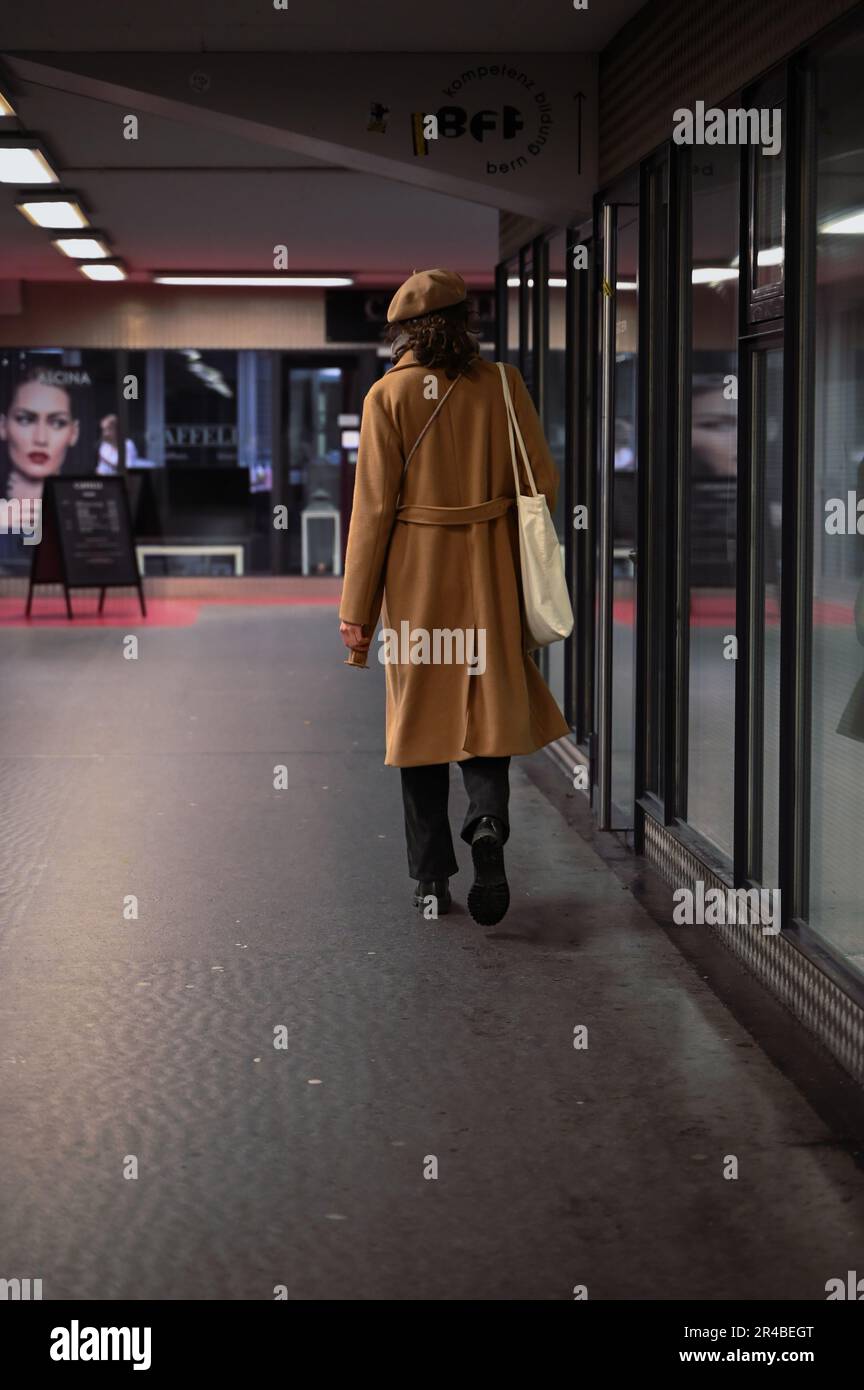 A lone figure in a brown coat strides past a window, with a determined look on their face, in a modern urban setting Stock Photo