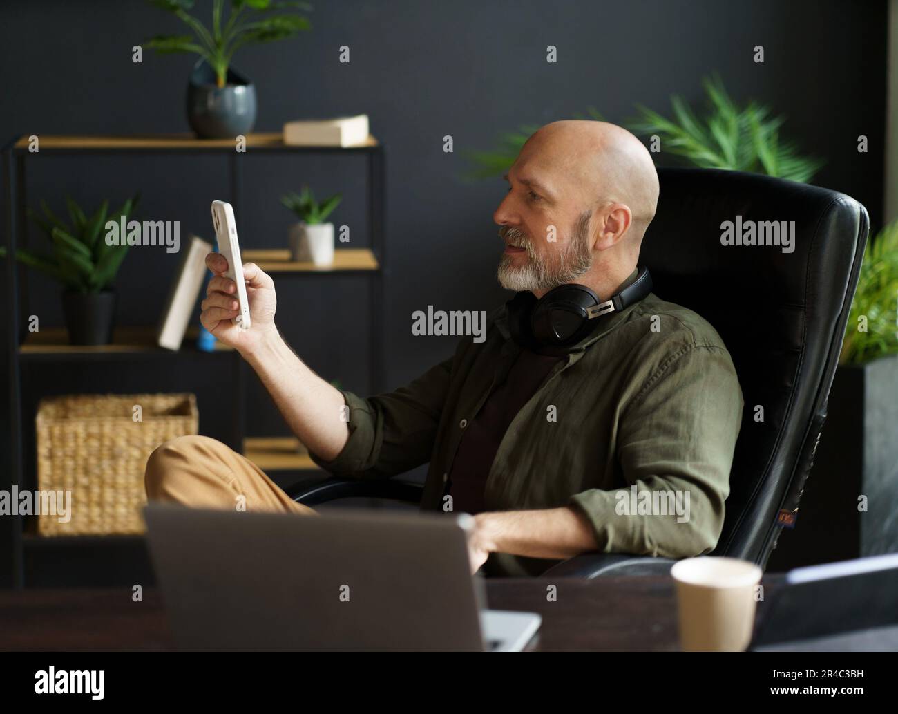 Smart and tech-savvy mid-aged man sits in chair at desk, engrossed in screen of phone. With bald head, silver beard, and air of handsomeness, multitasks, balancing phone, laptop, and cup of coffee. High quality photo Stock Photo
