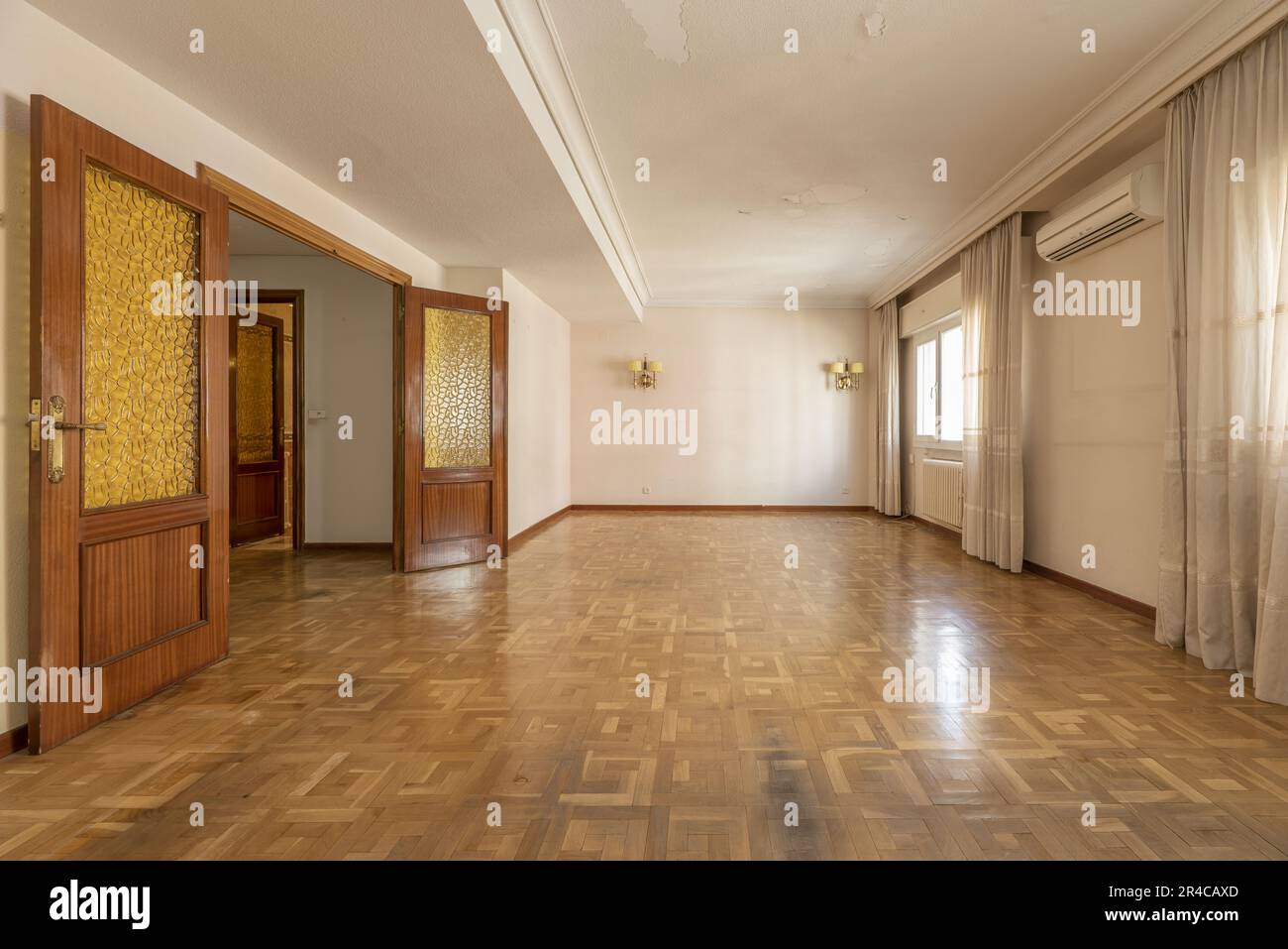 A large empty room with several windows with curtains, double sapelli doors with orange windows, oak parquet flooring, white aluminum radiators and wh Stock Photo