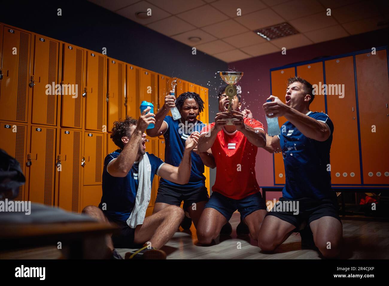 Group of young football players celebrating golden trophy together in the dressing room. Splashing water and shouting, feeling joyful. Stock Photo