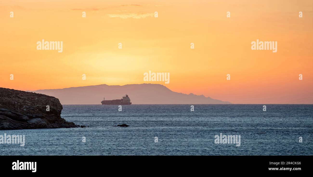 Sunset over Greek island as line ship leaves Cyclades. Sun colors the sky orange, yellow, hill silhouette. Calm vast blue Aegean sea. Space Stock Photo