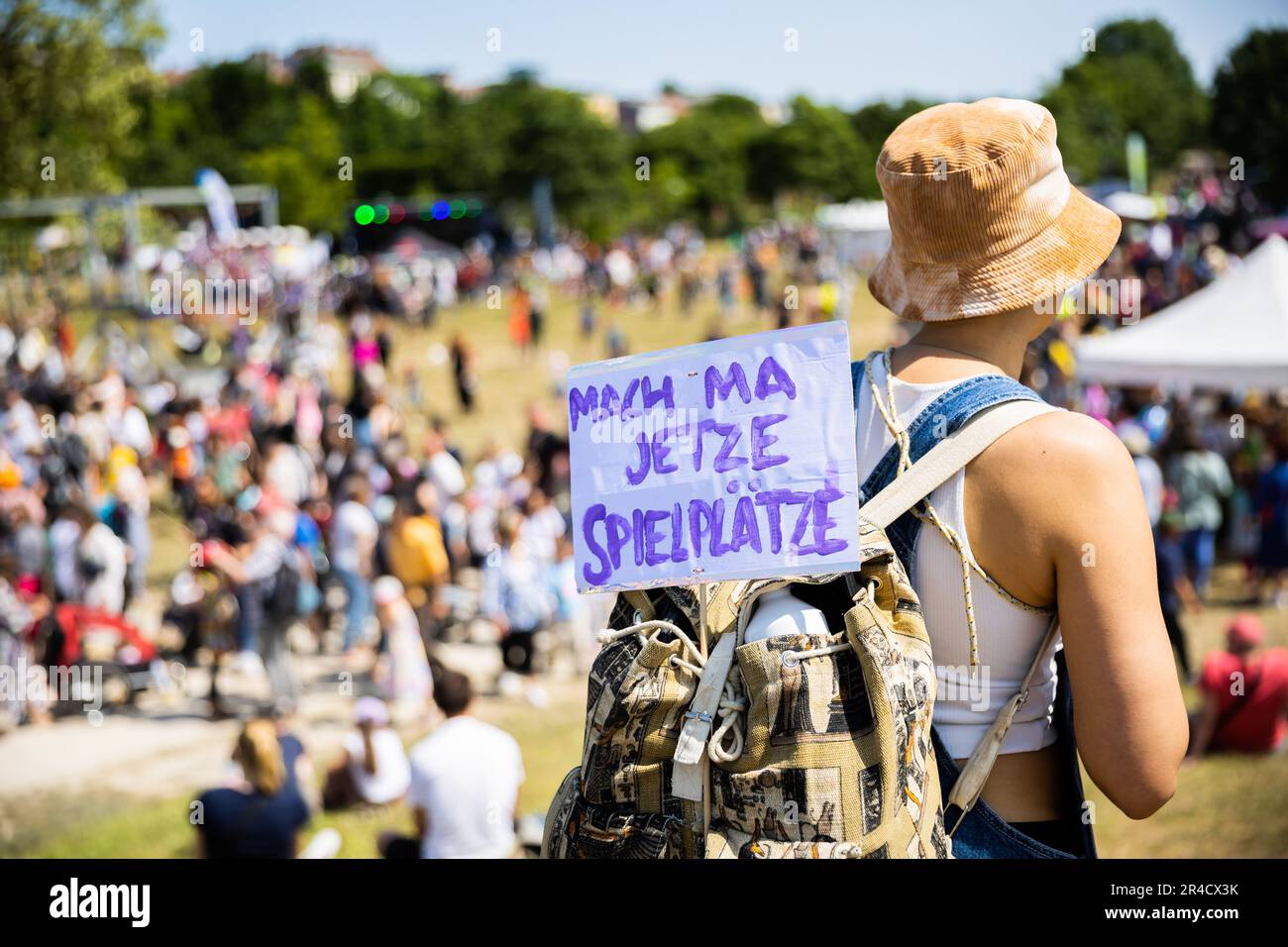 Berlin, Germany. 27th May, 2023. A woman carries a sign with the inscription 'Mach ma jetzte Spielplätze' on her backpack at the children's festival of the Children's Carnival of Cultures in Görlitzer Park. Around half a million people are expected to attend the 25th edition of the Carnival of Cultures in Berlin-Kreuzberg until Whit Monday. Credit: Christoph Soeder/dpa/Alamy Live News Stock Photo