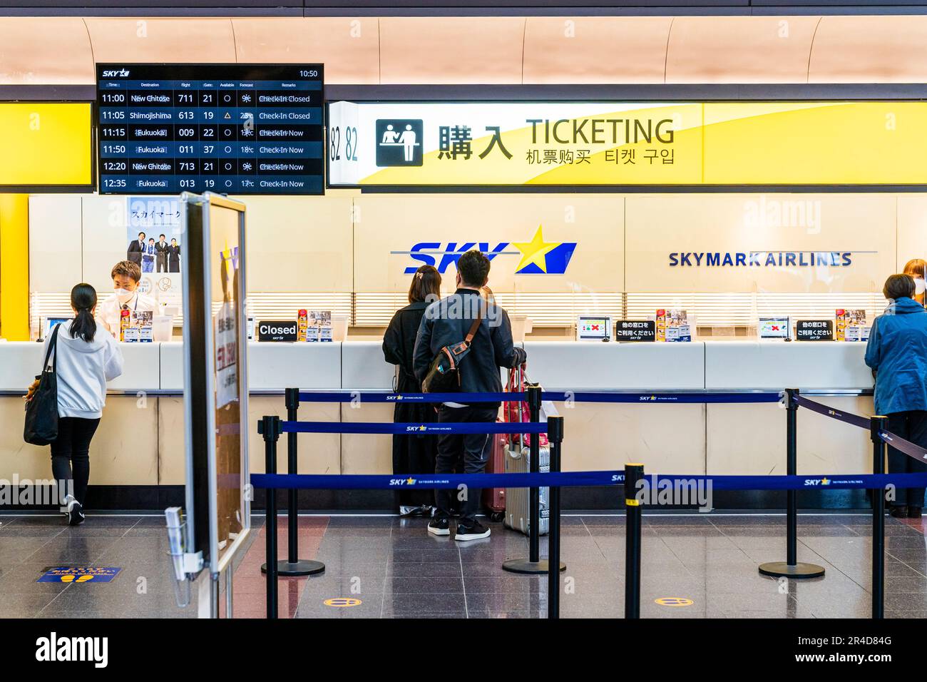 People at the Skymark Airlines ticketing counter at the North Wing of Terminal one, at Haneda Airport in Tokyo. Flight check-in display board above. Stock Photo