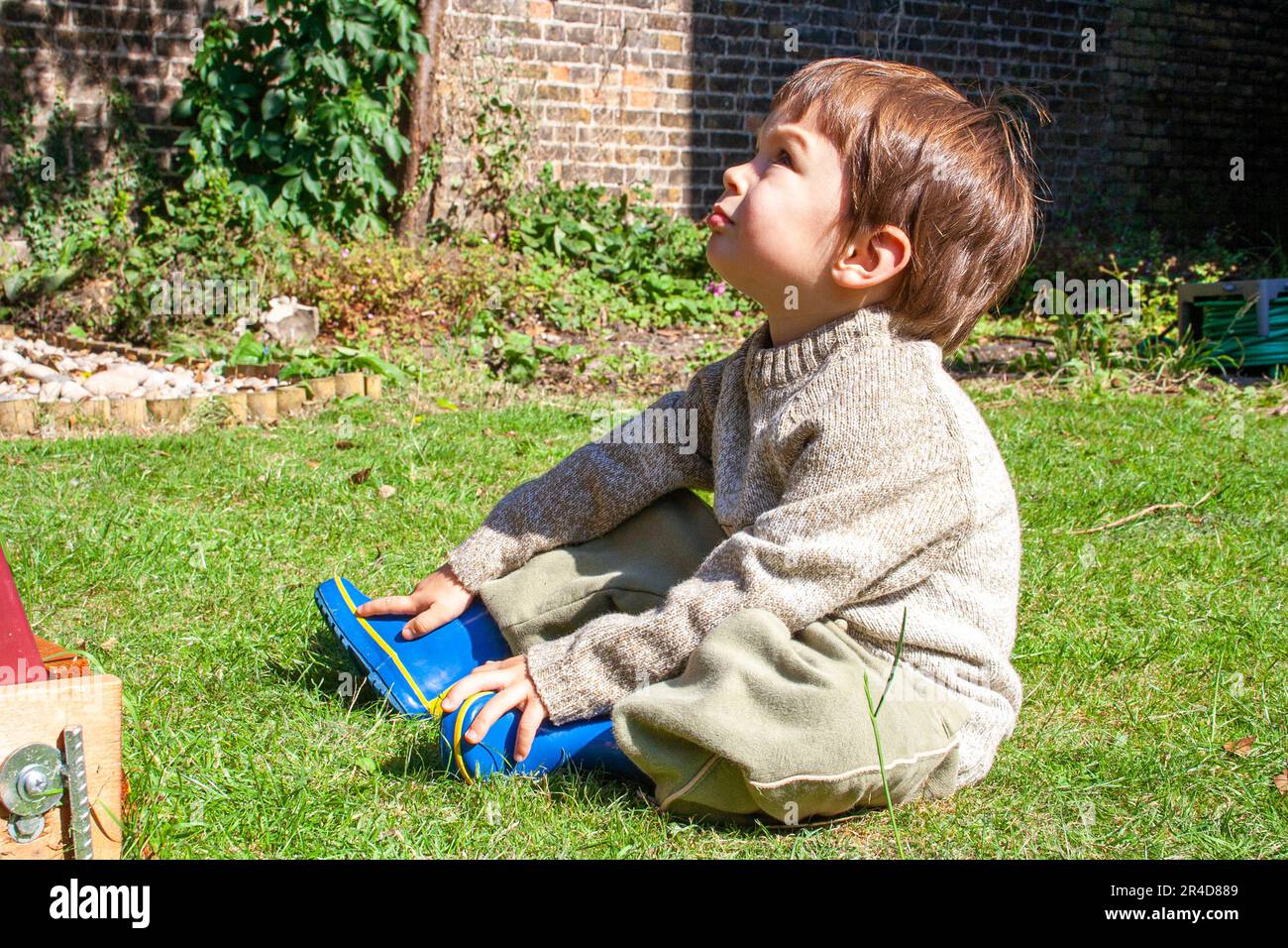 Young boy, child, 2 year old, sitting on grass in garden with hands on his boots, looking up at the sky in total contemplation. Bright sunshine. Stock Photo